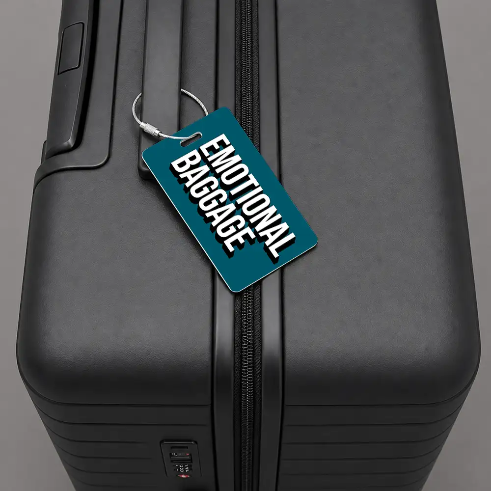 Close-up of a black hard-shell suitcase with a teal luggage tag reading 'EMOTIONAL BAGGAGE' attached to the zipper.