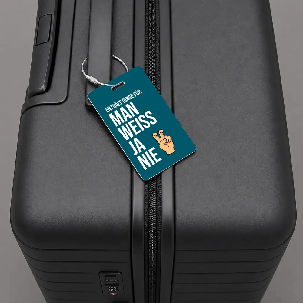 Close-up of a black hard-shell suitcase with a teal luggage tag that reads 'ENTHÄLT DINGE FÜR MAN WEISS JA NIE' and shows a hand emoji.
