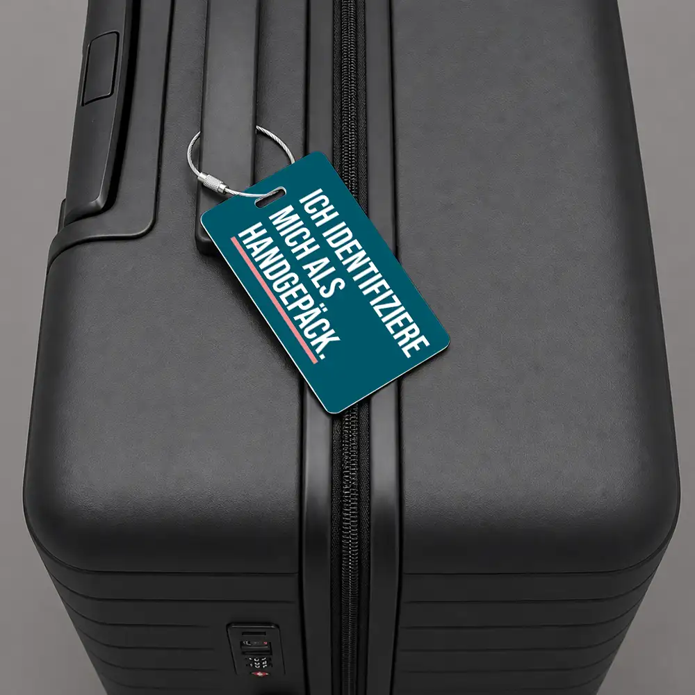 Close-up of a black hard-shell suitcase with a teal luggage tag hanging from the zipper, showing German text about hand luggage.