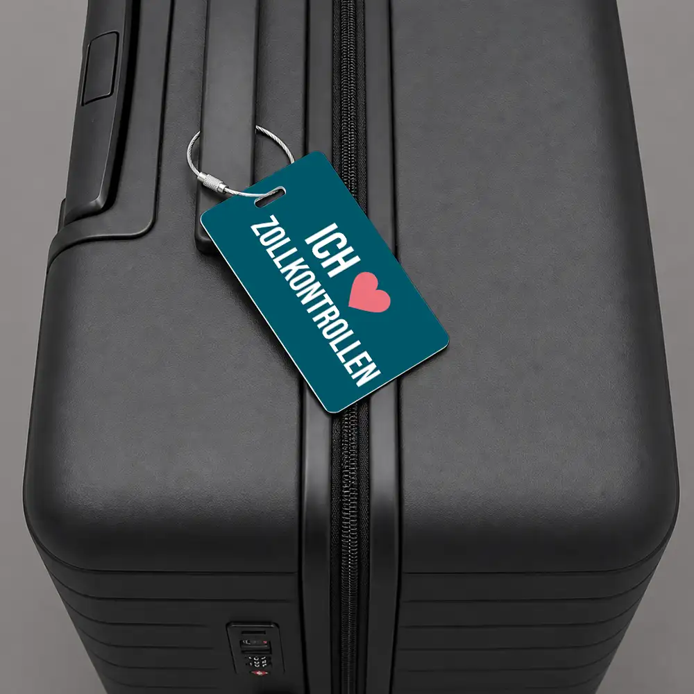 Close-up of a black hard-shell suitcase with a teal luggage tag attached to the zipper, showing the text 'ICH ♥ ZOLLKONTROLLEN' and a pink heart.