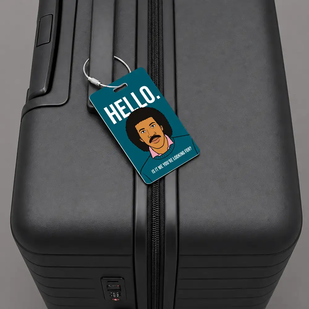 Close-up of a black suitcase with a teal luggage tag that says 'HELLO!' and features a cartoon man, attached to the zipper pull.