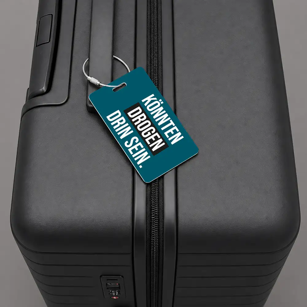 Black hard-shell suitcase with a teal luggage tag hanging from the zipper pull, the tag features bold white lettering.