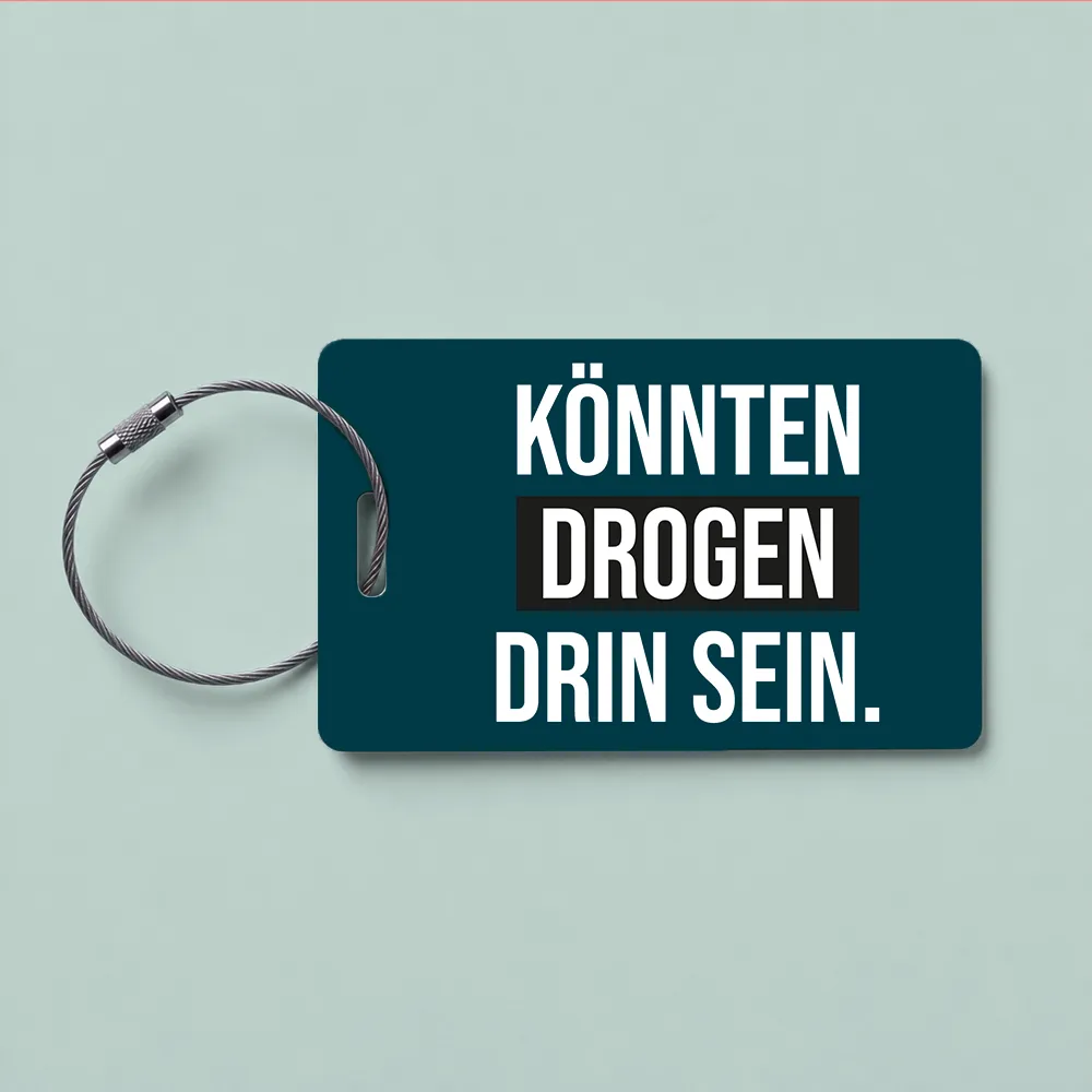 Tag with German phrase 'KÖNNTEN DROGEN SEIN' on a dark teal background, attached to a metal loop (drug-awareness message)