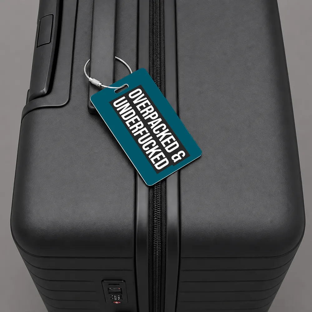 Close-up of a black hard-shell suitcase with a teal luggage tag attached to the zipper, displaying the text 'OVERBACKED & UNDERRUCKED'