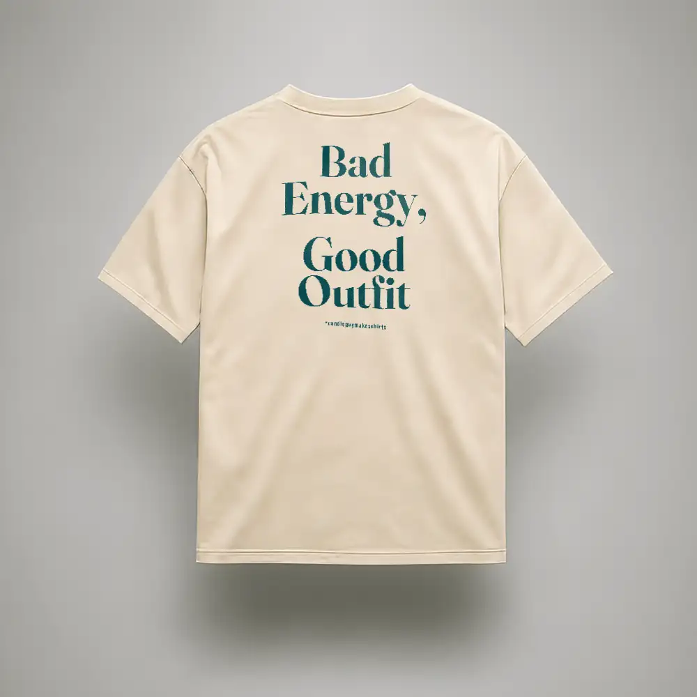 Beige short-sleeve T-shirt shown from the back with teal slogan: "Bad Energy, Good Outfit" and small hashtag text beneath.