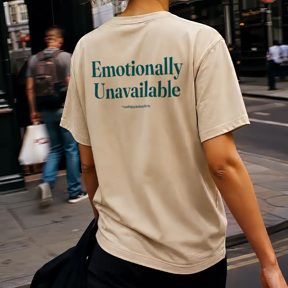 Back view of a person wearing a beige T-shirt with teal text reading 'Emotionally Unavailable' on a city street.