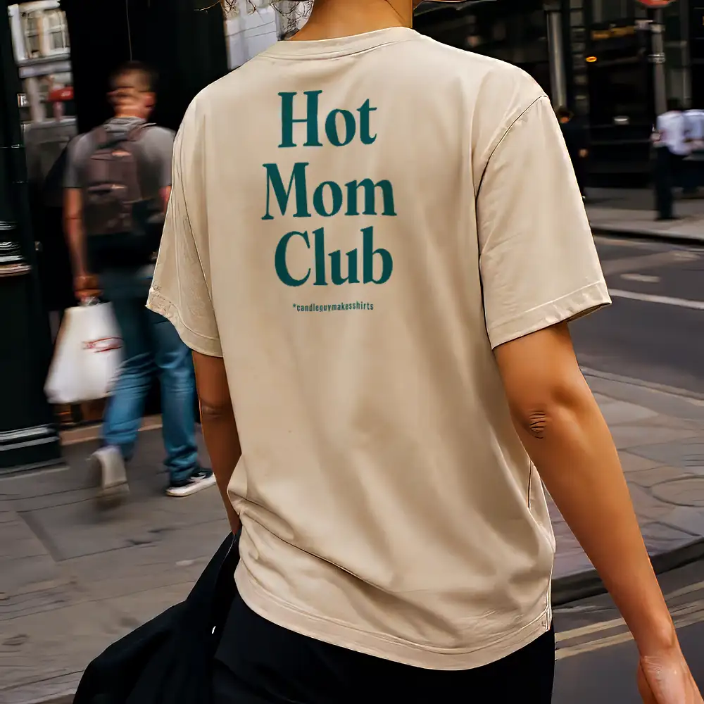 Back view of a person wearing a beige T-shirt on a city street; teal text on the back reads 'Hot Mom Club' with a small line of text beneath.