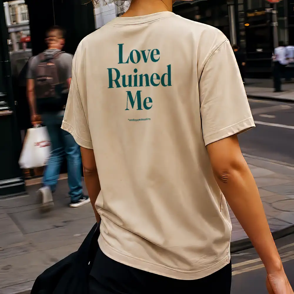 Person walking on a city street, seen from behind, wearing a beige T-shirt with teal text 'Love Ruined Me' on the back.