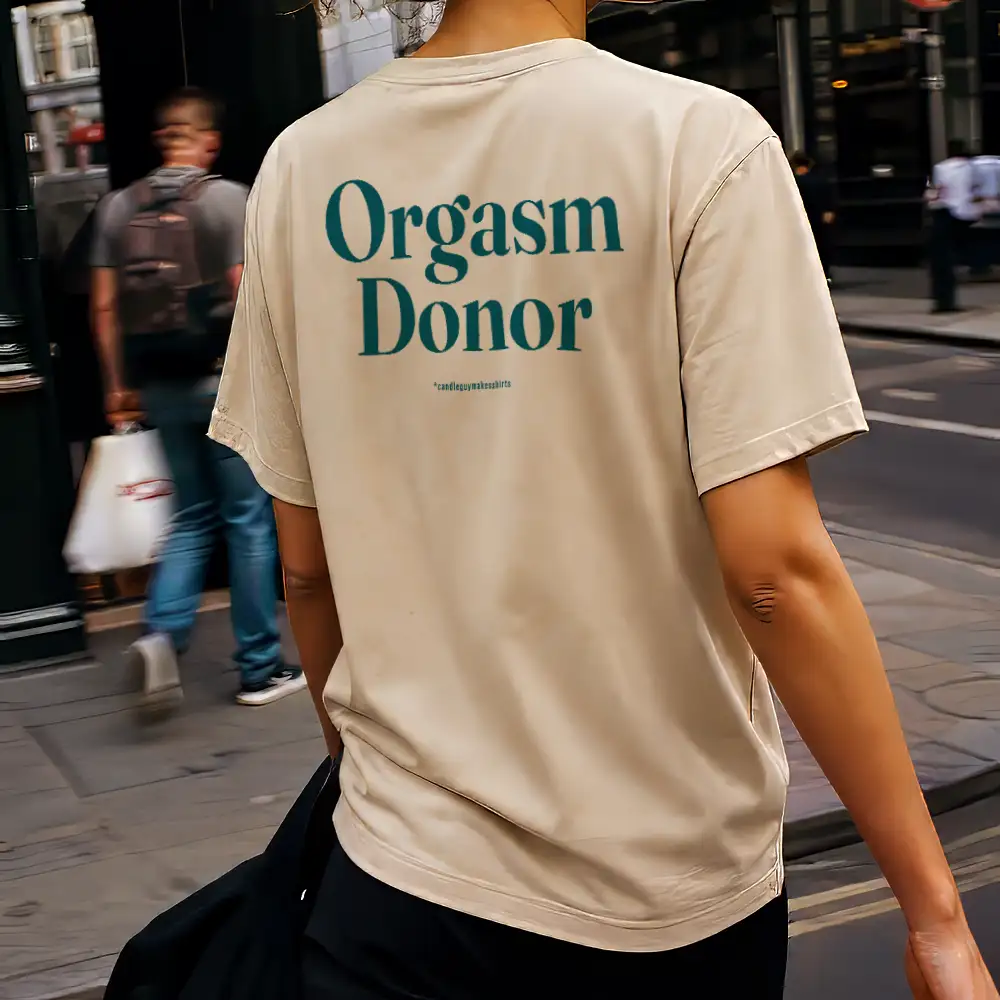 Person walking away on a city sidewalk wearing a beige T-shirt with teal text on the back reading 'Orgasm Donor'.